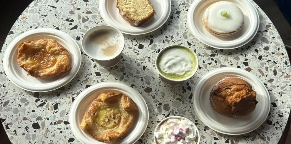 Overhead view of a selection of café treats and hot drinks arranged on a speckled terrazzo table. The spread includes pastries, a slice of loaf cake, an iced doughnut and a muffin, served on white plates. Several hot drinks are visible, including coffees, a green matcha-style drink with foam, and a hot chocolate topped with mini marshmallows. Natural light highlights the inviting café setting.