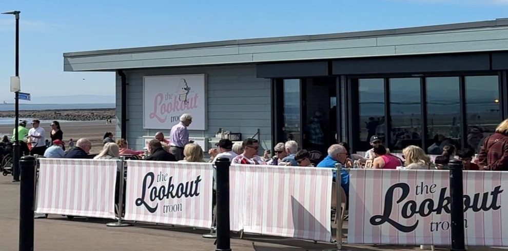 Exterior view of The Lookout Troon beachfront café on Troon Promenade on a sunny day. Customers are seated in the outdoor dining area behind branded windbreak panels featuring The Lookout Troon logo. The modern café building overlooks the beach and sea, with large windows reflecting the coastal views. The busy terrace highlights the café's relaxed, family-friendly atmosphere by South Beach.