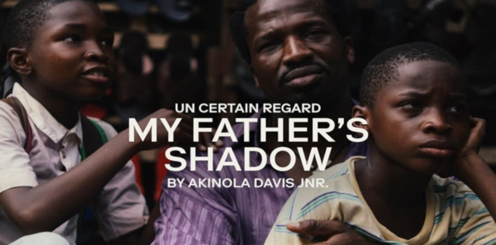 Promotional image for My Father's Shadow. Three people are shown seated close together in an outdoor setting. A central figure wearing a purple striped shirt sits between two younger figures, one on the left in a light-coloured collared shirt and one on the right in a striped T-shirt. The background is softly blurred, drawing attention to the group in the foreground. Large white text overlays the centre of the image, reading: “UN CERTAIN REGARD”, “MY FATHER'S SHADOW”, and “BY AKINOLA DAVIS JR.” The image uses warm, muted tones and a close, intimate composition, emphasizing the connection between the three figures and reflecting the film's themes of family, memory and shared experience.