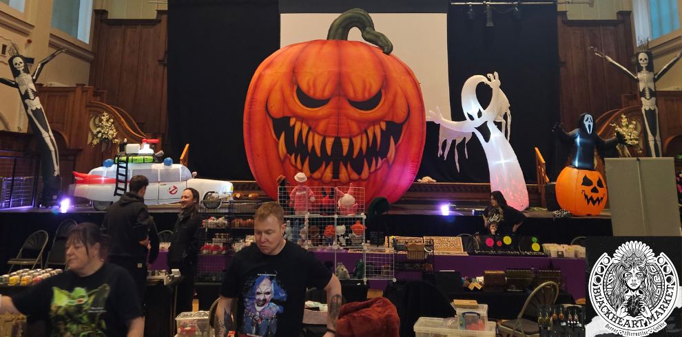Wide view of a Halloween-themed market inside a large hall. At the front of a stage is a giant inflatable jack-o'-lantern with a menacing face, surrounded by Halloween decorations including skeletons, ghosts, and pumpkin displays. A replica Ghostbusters vehicle sits to one side of the stage. Market stalls filled with horror, gothic, and alternative merchandise are set up below, with visitors browsing throughout the venue. A Blackheart Market logo watermark appears in the bottom right corner.