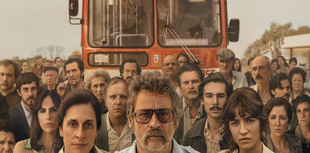 Film poster featuring a large group of people standing closely together in the foreground, facing the camera. At the centre is a person wearing large glasses, surrounded by individuals of various ages. Behind the group is the front of a red city bus, which dominates the background and serves as the focal point of the composition. The image uses warm, earthy tones and period-style clothing, suggesting a historical setting. Buildings and trees are faintly visible in the distance on either side of the bus, creating the impression of a neighbourhood street scene.