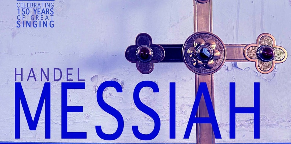 Promotional poster for Handel's Messiah. Large blue text reading “HANDEL MESSIAH” appears across the lower half of the image. A decorative metal church cross with ornate circular details is positioned prominently in the centre-right against a pale, textured wall. In the top left corner, smaller text reads “Celebrating 150 Years of Great Singing.” The image uses cool blue and purple tones, creating a dramatic and reverent atmosphere.