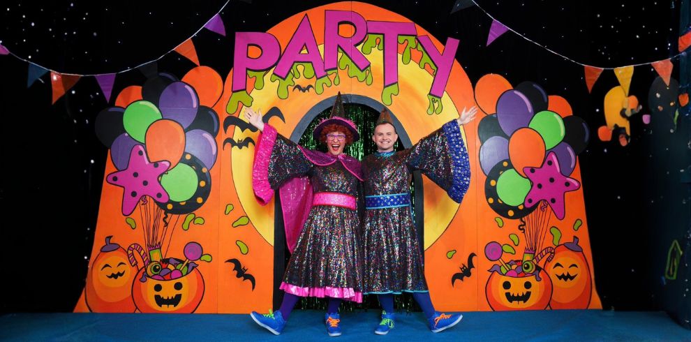 Two performers in colourful Halloween-themed costumes stand beneath a large orange archway spelling “PARTY” on a decorated stage. The stage is surrounded by clusters of orange, purple, green and black balloons, with pumpkin buckets, bats, bunting and Halloween decorations throughout. The performers have their arms raised and wear matching sparkly capes and bright trainers. The backdrop features a starry night sky, creating a fun and festive setting for The McDougalls Halloween Party Show.