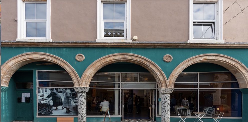 Front view of a historic shopfront in Ayr with three large stone arches framing wide windows and a central entrance. The building is painted teal at street level, with upper-storey sash windows above. A large black-and-white photograph is displayed in the left window, while people can be seen gathering inside the building. Outdoor café-style tables and chairs sit in front of the right-hand window, and the façade combines traditional stone detailing with a newly activated community space.