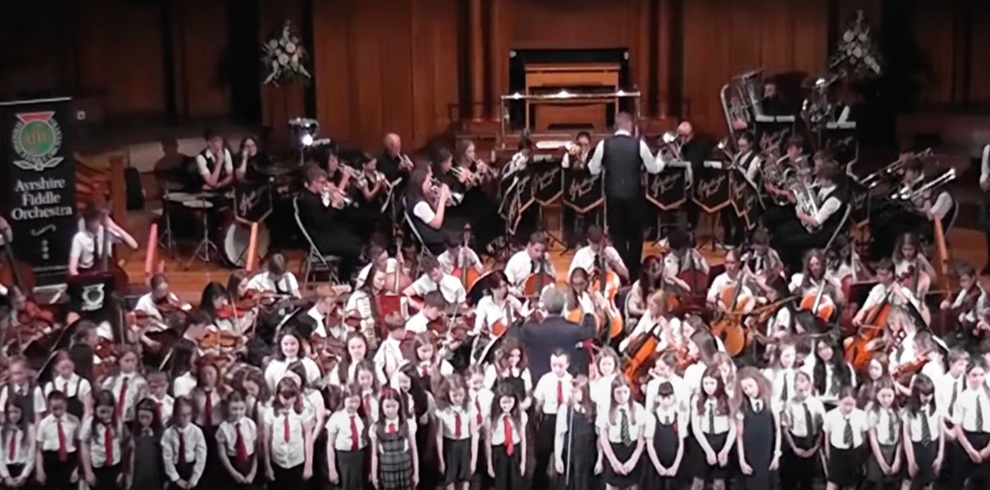 A large musical performance taking place on a concert hall stage. In the foreground, a children's choir dressed in white shirts and dark skirts or trousers stands in rows. Behind them, a large orchestra made up of string, brass and percussion musicians performs under the direction of conductors. A banner for the Ayrshire Fiddle Orchestra is visible on the left side of the stage. The venue features wood-panelled walls, floral arrangements and tiered seating for the performers.