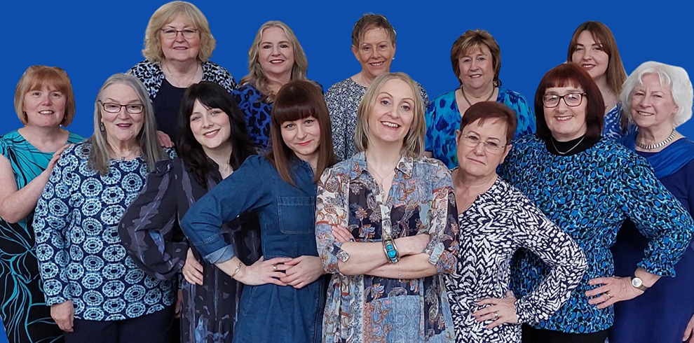 Group promotional photograph of Coast Harmony, a vocal ensemble featuring twelve members arranged in three rows against a solid blue background. The group is dressed in coordinated blue, black and patterned clothing, with several members standing and others positioned in front. The image has a friendly, professional appearance and is suitable for promoting a live music performance.