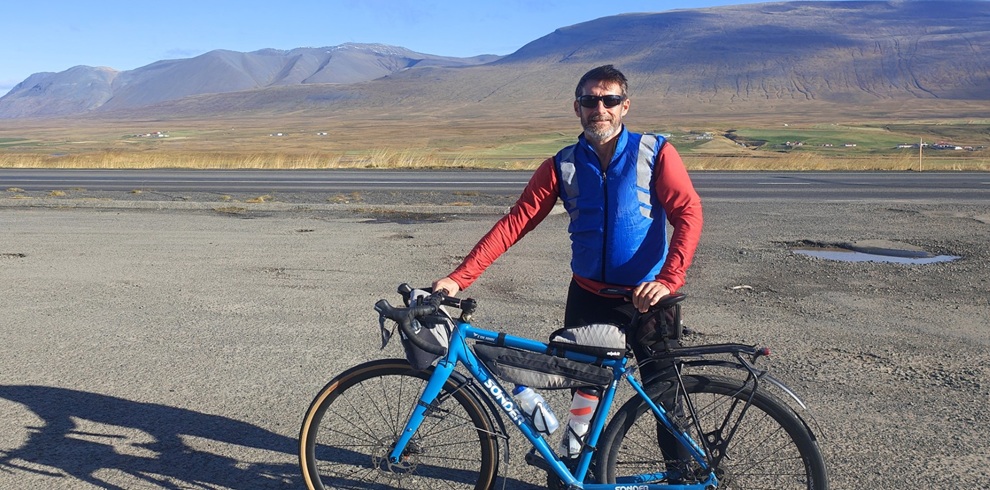 A person standing beside a blue touring bicycle on a wide gravel roadside in a remote countryside setting. The bicycle is fitted with storage bags and water bottles. Rolling brown hills and mountains stretch across the background under a clear blue sky, with long shadows cast by the low sunlight. A few scattered buildings can be seen in the distance.