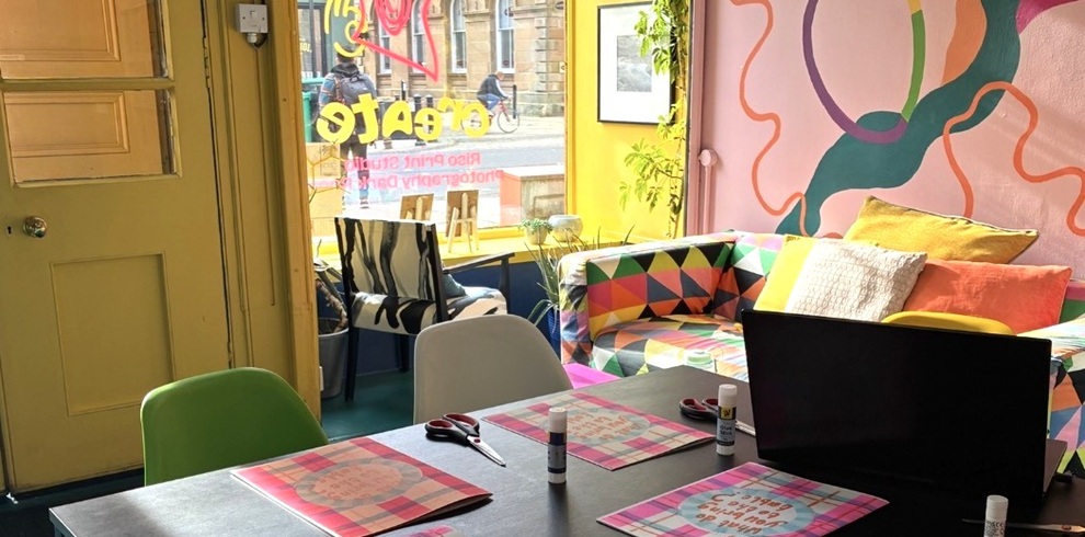 Colourful interior of Narture’s Create studio, featuring a workspace set up for a printmaking activity. In the foreground, a large table holds scissors, glue sticks and printed craft materials, alongside a computer monitor. Brightly coloured chairs surround the table. In the background, a geometric-patterned sofa sits against a mural-painted wall in shades of pink, orange, green and purple. Natural light streams through the front window, where the reversed word “Create” and additional studio signage are visible from inside. Plants, framed artwork and vibrant décor create a welcoming and creative atmosphere.