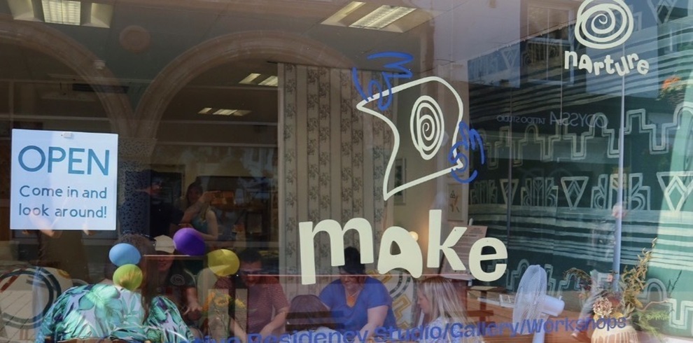 Street-facing window of Narture’s Make creative residency studio and gallery in Ayr. The large glass frontage displays the word “make” and the Narture logo in white vinyl lettering, alongside an “OPEN – Come in and look around!” sign. Through the window, several people are gathered around tables inside the bright studio space, with colourful balloons and artwork visible. Reflections of the historic arched building opposite can be seen in the glass, highlighting the venue’s role as an active and welcoming community creative hub.