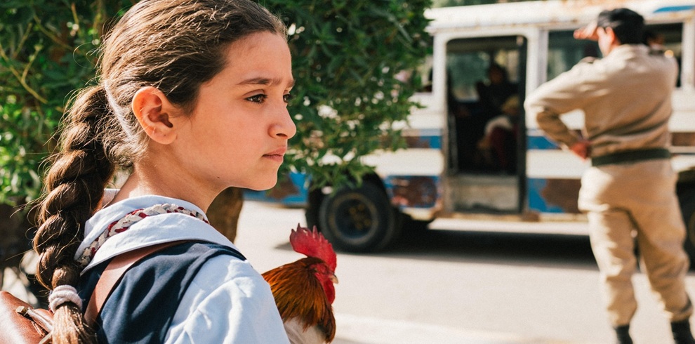 Close-up promotional still showing a young child with long braided hair standing beside a road and holding a chicken. The child wears a light-blue collared shirt under a dark garment with patterned trim. In the background, a white bus with blue stripes is parked with its door open, while a uniformed figure stands nearby. Trees and greenery frame the scene, and the image is lit by warm natural sunlight.