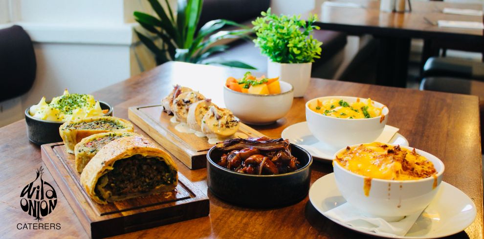 Selection of catered hot dishes arranged on a wooden table in a restaurant setting, including a sliced savory pastry roll, stuffed chicken with creamy sauce, caramelized onions, potato dishes, and vegetables served in bowls and dishes. Potted plants and dining tables are visible in the background. The Wild Onion Caterers logo appears in the lower-left corner.