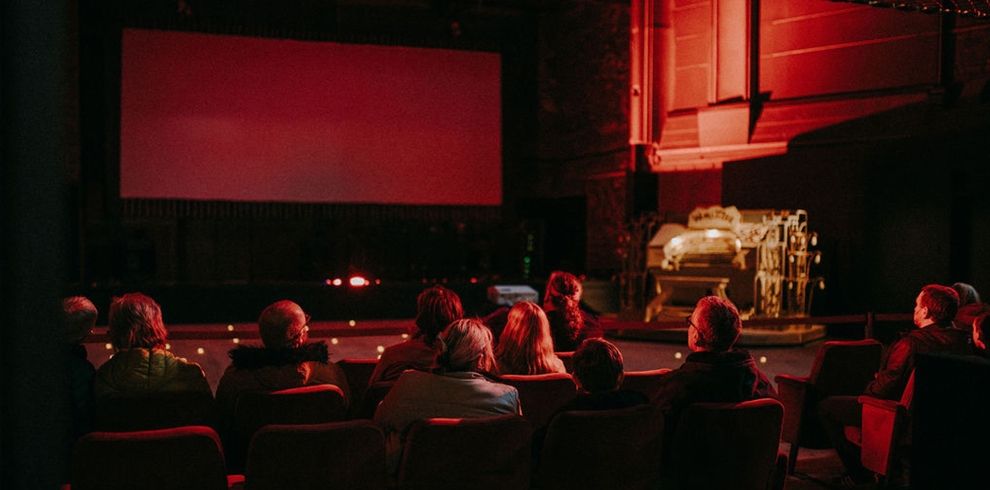 Audience members sit in the Broadway Cinema auditorium watching film clips on a large screen. The historic theatre is illuminated with warm red lighting, with rows of seats in the foreground and vintage cinema equipment visible beside the stage.
