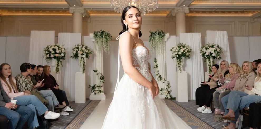 The image shows bridal catwalk scenes at an elegant wedding exhibition venue, featuring wedding dresses on a runway surrounded by floral displays, chandeliers and seated visitors watching the fashion showcase. The setting highlights wedding inspiration, bridal fashion and wedding planning services available at the event.