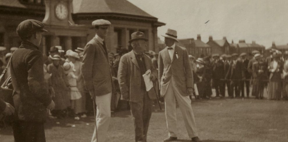 An old sepia photograph of 3 people golfing and 1 boy caddying in front of a crowd outside a clubhouse.