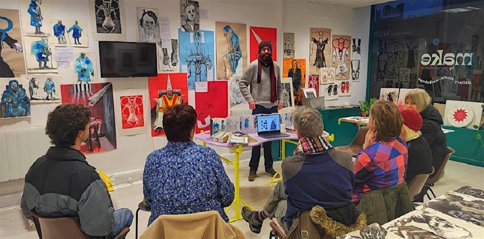 A photograph of an art workshop or artist talk taking place in a gallery space. A bearded man wearing a gray sweater, red scarf, and beanie stands at the front, speaking to an audience seated in chairs. The walls are filled with expressive artwork, including ink and watercolor portraits, figurative sketches, and abstract compositions in blue, red, and black tones. A television screen and a laptop on the table display digital images related to the presentation. The audience, dressed in winter clothing, attentively listens. The gallery has a creative and intimate atmosphere, with artwork arranged in a dynamic and engaging way. A glass window with the word "Make" in white letters is visible in the background.