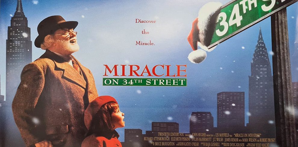 A still from the movie Miracle on 34th Street featuring the two main characters: Kris Kringle, an older man with a full white beard wearing a Santa Claus outfit, and Susan Walker, a young girl with curly hair wearing a winter coat and hat, smiling up at him.