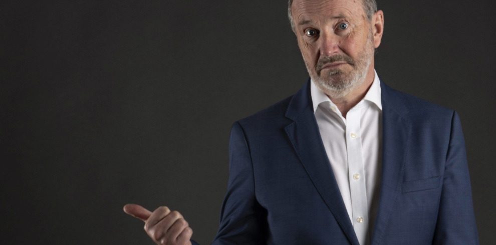 Comedian Fred MacAulay standing against a dark background indoors.