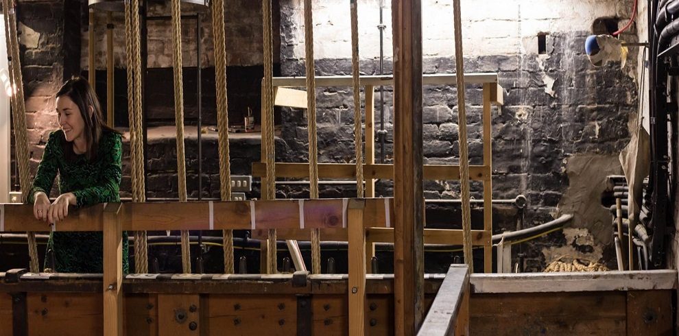 The image shows the backstage area of the Gaiey Theatre, specifically the space above the curtains behind the stage. This area is characterized by a network of catwalks, ropes, and pulleys used for managing the theater's curtains and stage props. The lighting is dim, with beams and structures visible, creating an industrial and functional atmosphere. The setup indicates the technical side of theater production, with various equipment and safety rails in place.