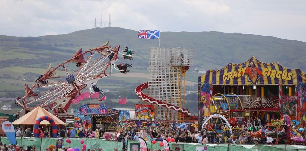 Fairground rounds at busy festival with hills in the background.