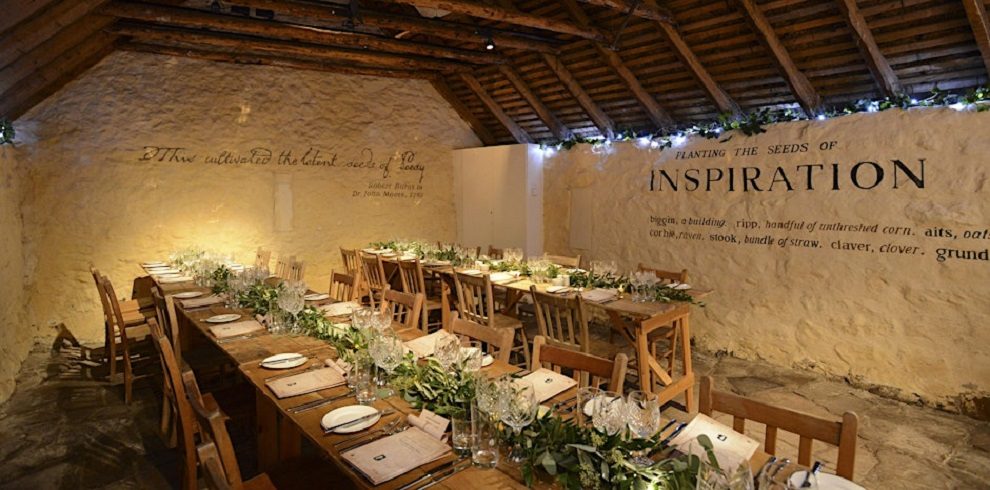 The image shows the interior of a rustic, stone-walled room, likely part of Burns Cottage. The space features a high, sloping ceiling with exposed wooden beams. Wooden tables and chairs are neatly arranged in rows, set for a formal meal. The tables are decorated with greenery, white flowers, and neatly arranged plates, glasses, and utensils. Text is painted on the stone walls, including phrases like "Planting the seeds of INSPIRATION" and quotes referencing Robert Burns and agriculture. Soft lighting and garlands of lights along the beams create a warm, inviting ambiance.