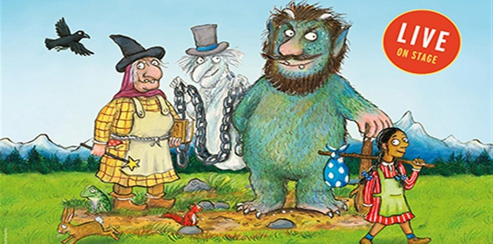 The image is an illustration promoting a live stage show called The Baddies. It features four main characters in a grassy, outdoor setting with mountains in the background. From left to right, there’s a witch wearing a yellow dress with a red scarf, holding a brown spellbook, and a crow flying beside her. Next to her is a ghostly figure in a top hat and chains. A large, green troll with shaggy fur and a club stands to the right, towering over a young girl. The girl, dressed in bright, colorful clothing, carries a bindle on a stick, as if she's on an adventure. In the top right corner, there's a red circle with the words "LIVE ON STAGE.