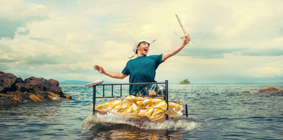 A picture of a person floating on what looks like a bed in the sea. Their left arm is stretched out, holding a sword in the air.